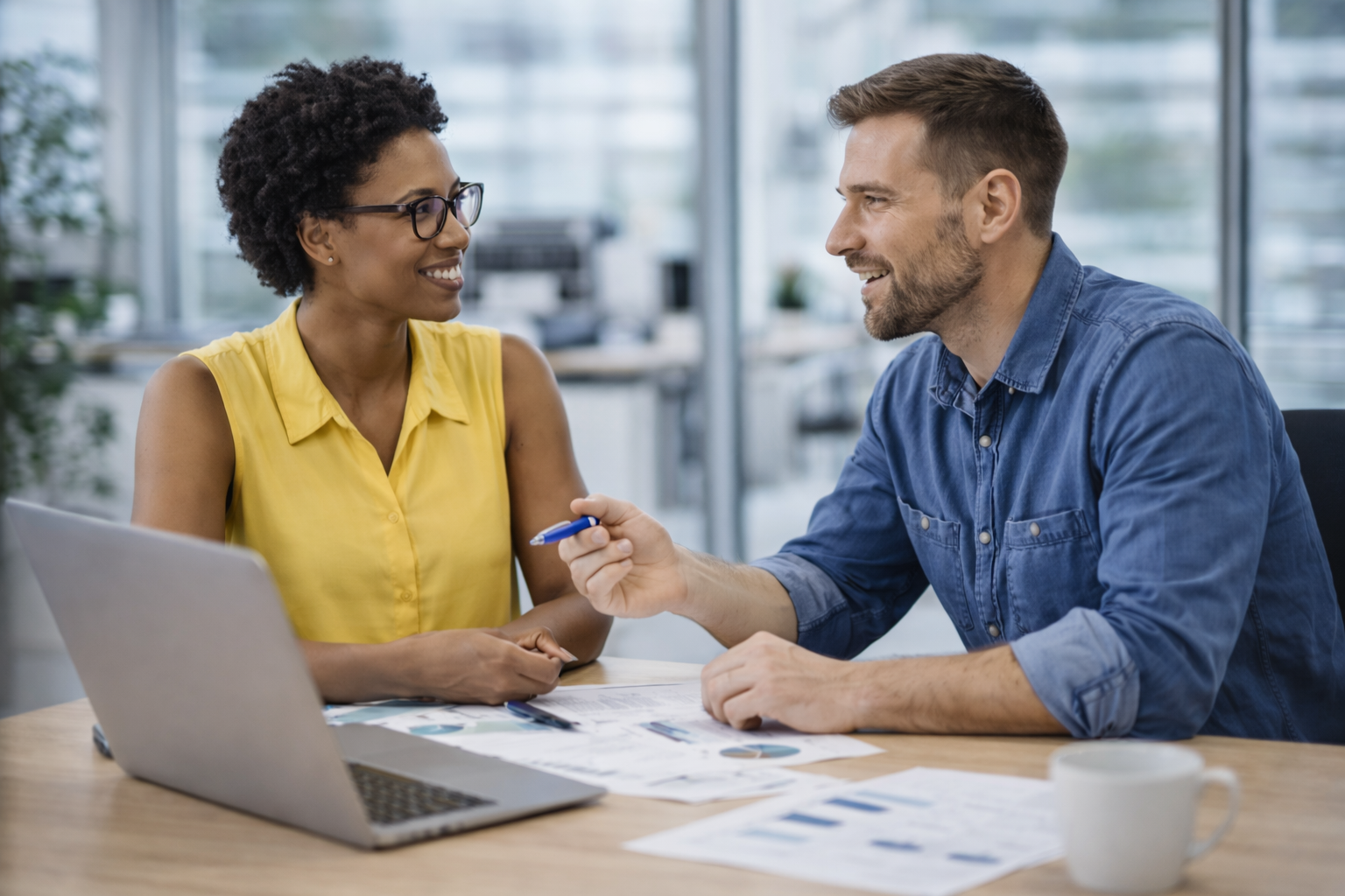 Ein Mann und eine Frau sitzen an einem Schreibtisch mit Laptop und Unterlagen und diskutieren in einer hellen Büroumgebung über Weiterbildung.
